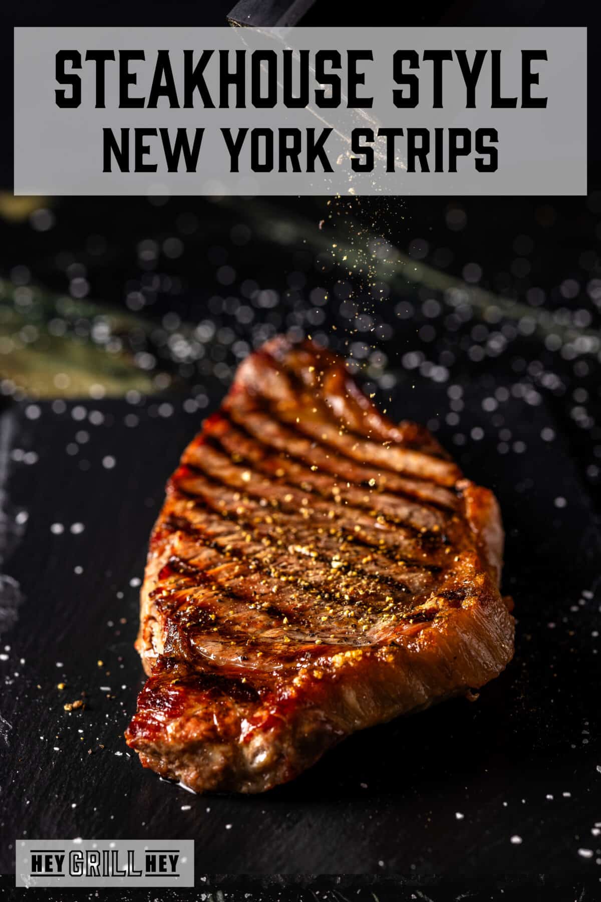 A grilled steak on a black surface being sprinkles with seasoning. The text overlay reads "Steakhouse Style New York Strips" at the top, and "Hey Grill Hey" at the bottom.