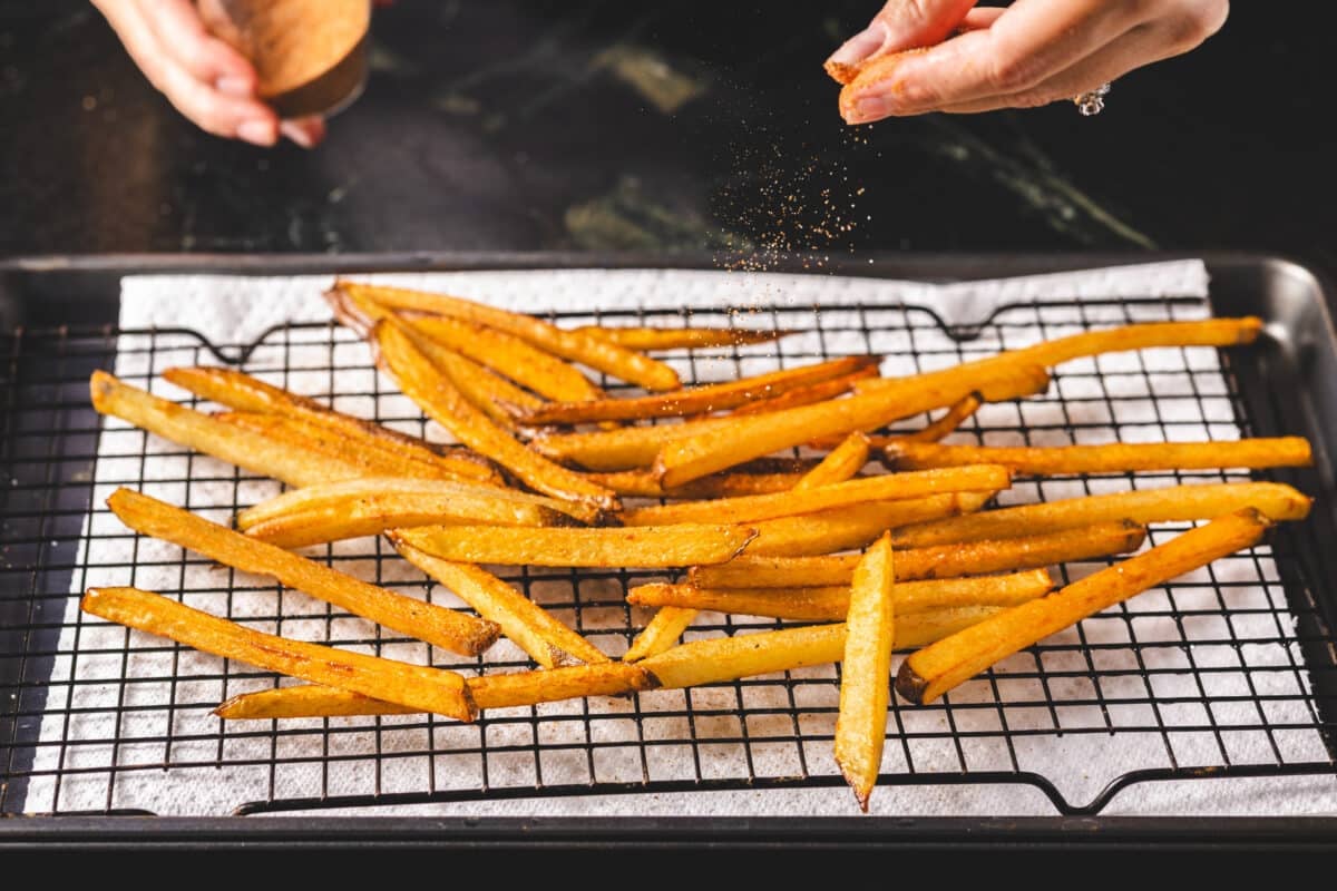 A chef's hands sprinkling seasoning onto cooked fries on a wire rack.
