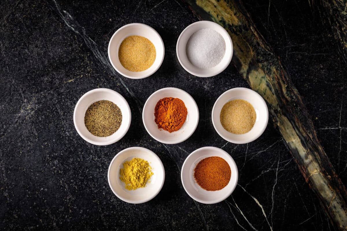 Seven bowls of various spices arranged in rows of two, three, and two, on a black marble surface.