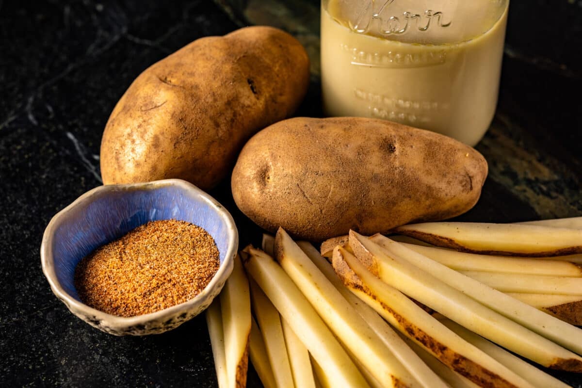 Two raw potatoes, a julienned potato, a jar of tallow, and a bowl of spice rub on a black countertop.