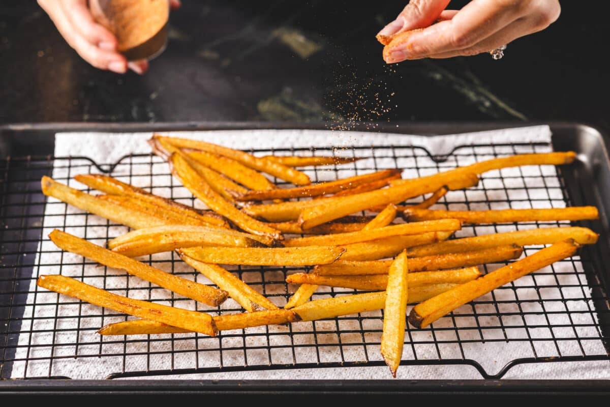 A chef's hands sprinkling seasoning onto fresh fries on an elevated baking sheet lined with parchment paper.