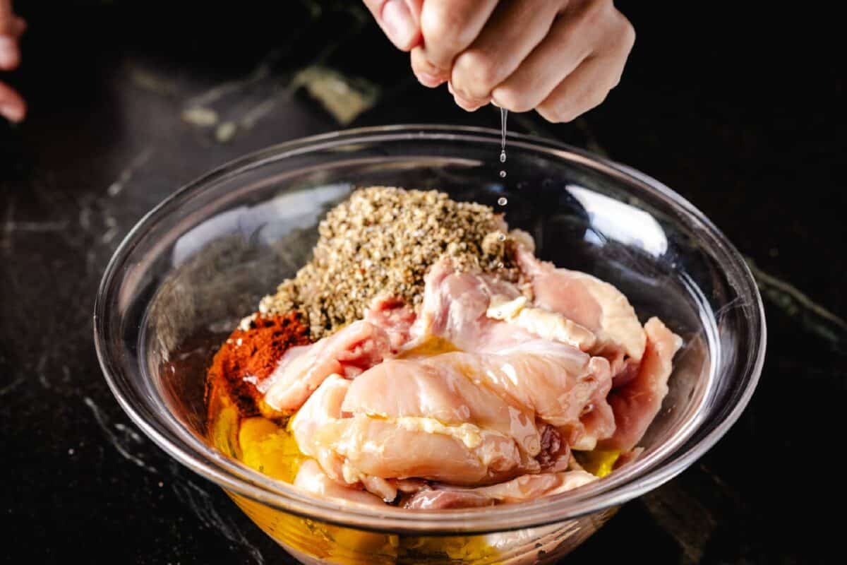 A chef's hand squeezing fresh lemon juice into a clear bowl of raw chicken and various seasonings.