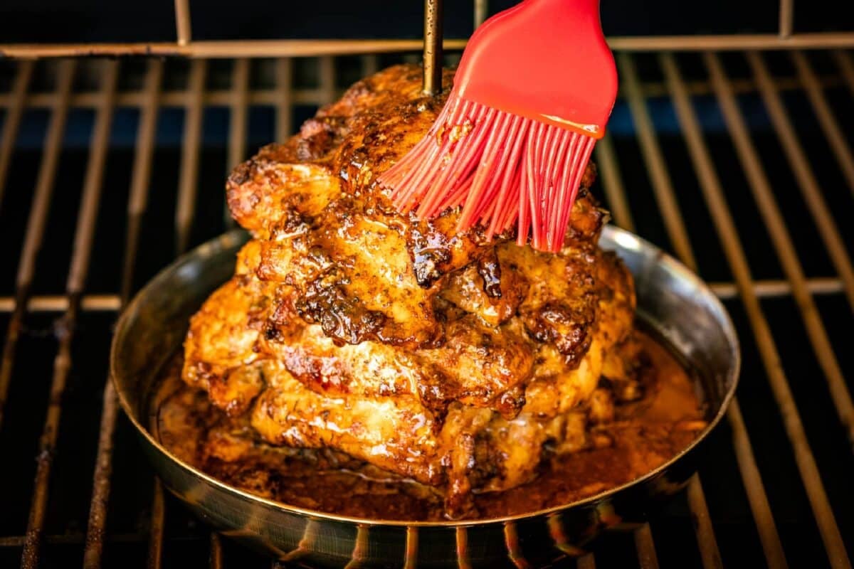 A red basting brush glazing chicken on a trompo sitting on the grates of a smoker.
