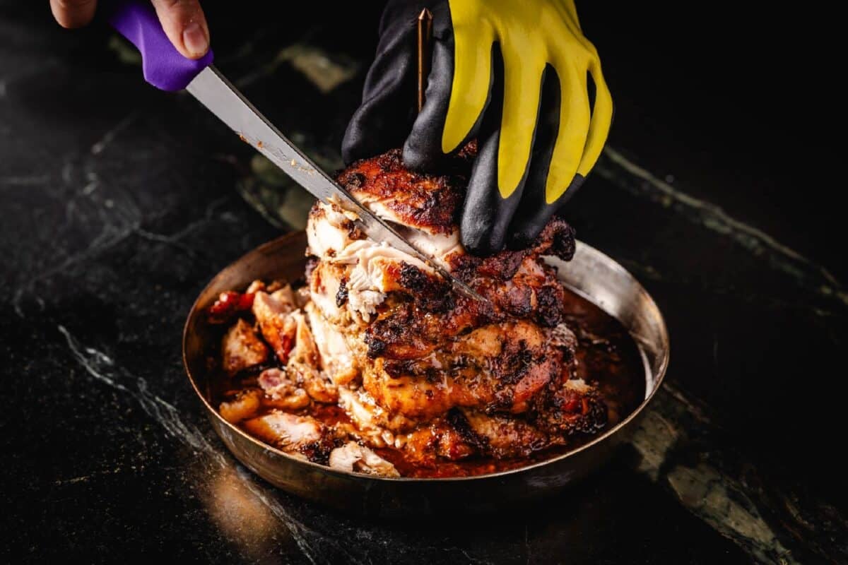 A gloved hand holding smoked chicken gyro meat in place as an ungloved hand slices the chicken with a knife.