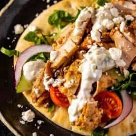 An open-faced smoked chicken gyro topped with chopped chicken and diced veggies on a black plate.