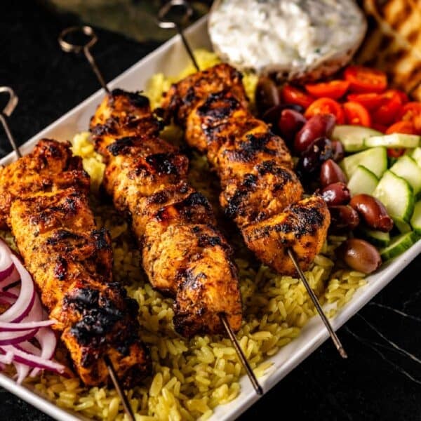 A platter of skewered chicken on a bed of rice with a cup of tzatziki sauce and fresh tomato and cucumber as a garnish.