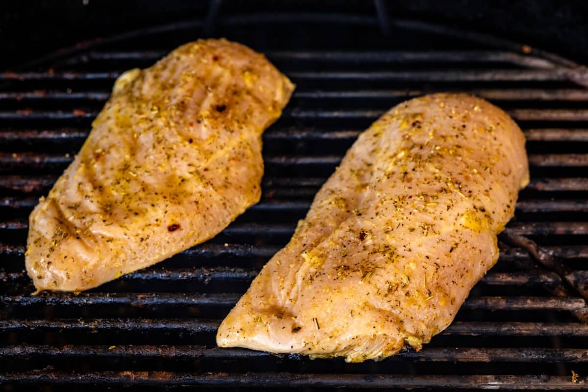 Two seasoned chicken breasts on the grates of a smoker.