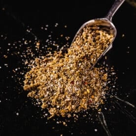 A pile of Greek seasoning spilling out from a metal scoop onto a black surface.
