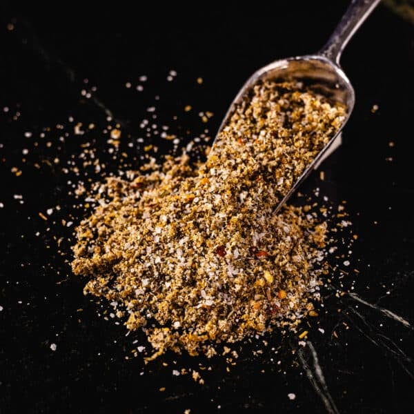 A pile of Greek seasoning spilling out from a metal scoop onto a black surface.