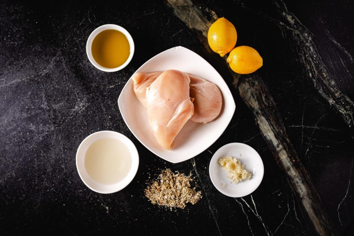 An overhead shot of a white bowl of raw chicken, smaller bowls of liquids and seasonings, and fresh lemons on a dark surface.