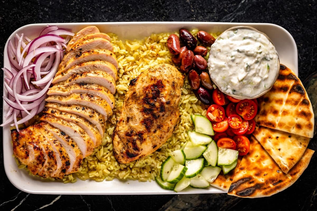 A white platter of sliced chicken and veggies, toasted pitas, and a bowl of tzatziki sauce.