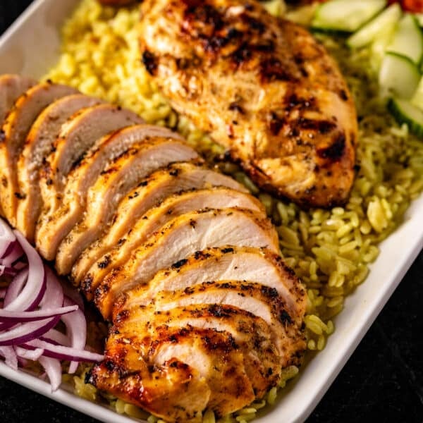 A white platter of sliced Greek-style chicken and veggies on a bed of rice.