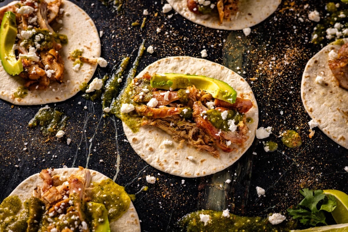 Several smoked chicken tacos topped with avocado and salsa verde on a marble surface.