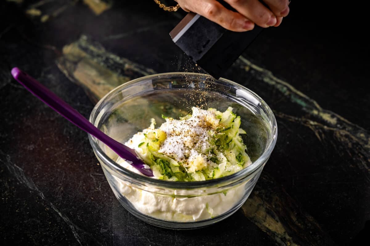 A chef's hand adding black pepper to a clear bowl filled with dressing. There is a purple spatula resting in the bowl.