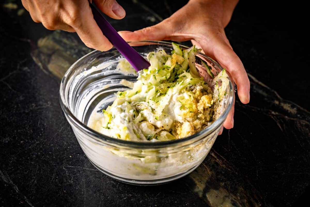 A chef's hand stirring a dressing in a clear bowl with a purple spatula.