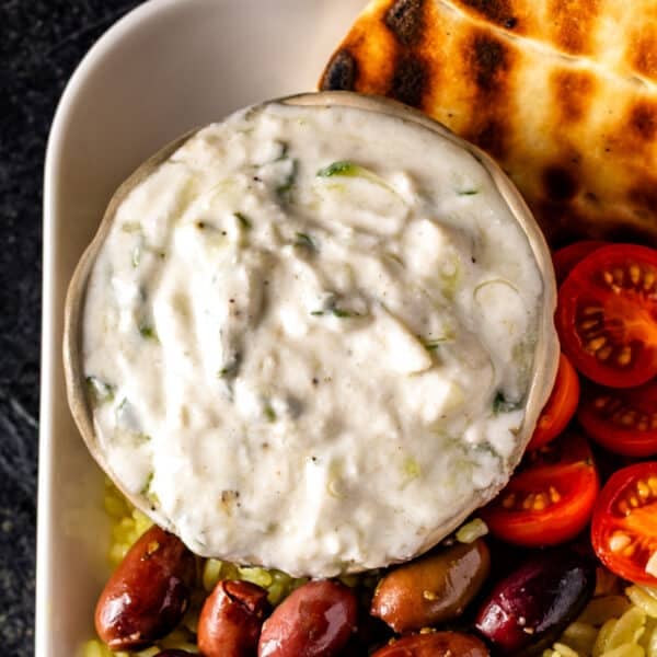 A closeup shot of a bowl of tzatziki sauce in the corner of platter with pita bread, olives, and cherry tomatoes.