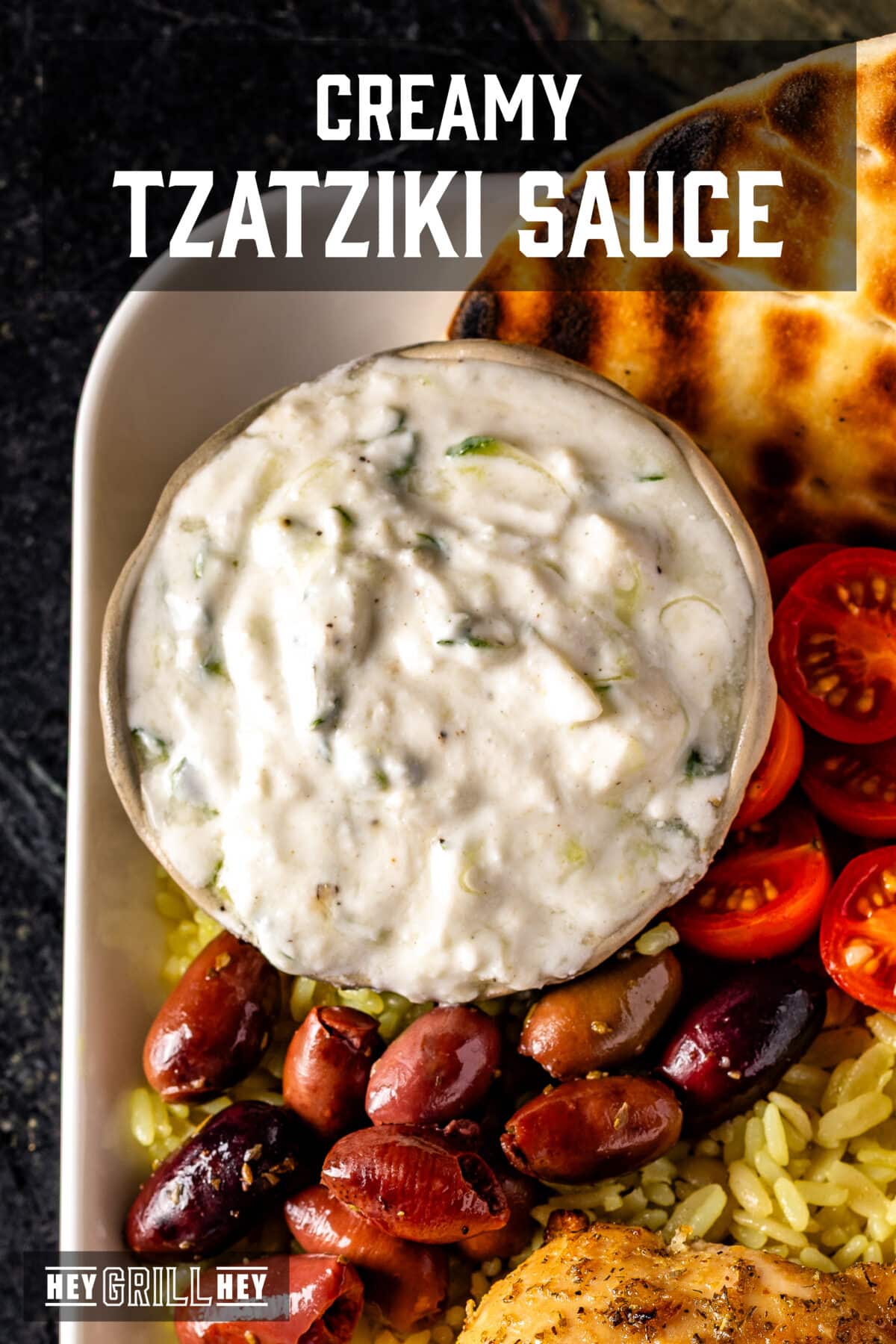 A closeup shot of a bowl of tzatziki sauce in the corner of platter with pita bread, olives, and cherry tomatoes. The text overlay reads "Creamy Tzatziki Sauce" at the top, and "Hey Grill Hey" at the bottom.