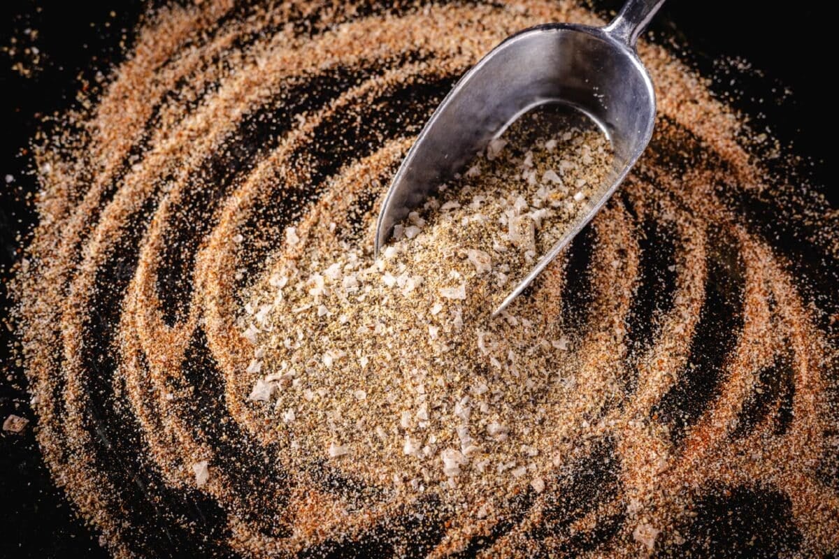 A metal scoop in a BBQ rub piled on a dark surface.