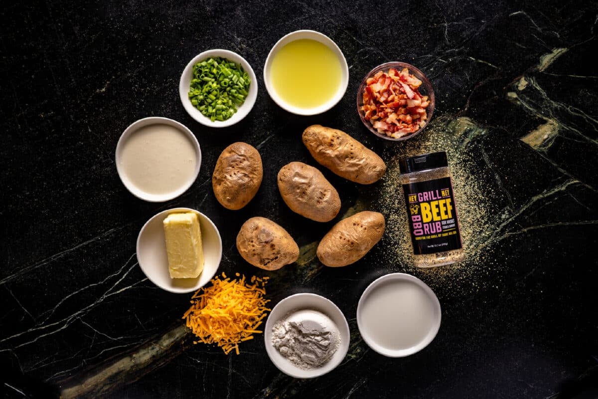 Five uncooked potatoes on a counter surrounded by bowls of liquids and chopped veggies, a pile of shredded cheese, and a bottle of Hey Grill Hey Beef Rub.