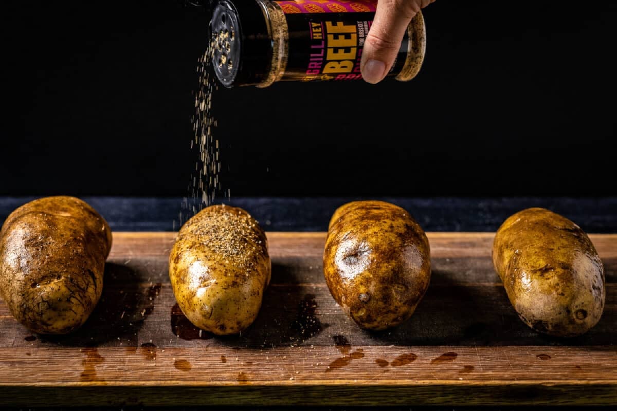 A chef's hand sprinkling Hey Grill Hey Beef Rub onto oiled potatoes resting on a wooden cutting board.