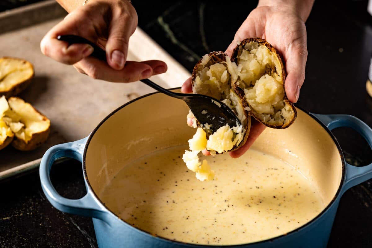A chef's hand scooping the inside of a potato into a large pot of liquid using a spoon.
