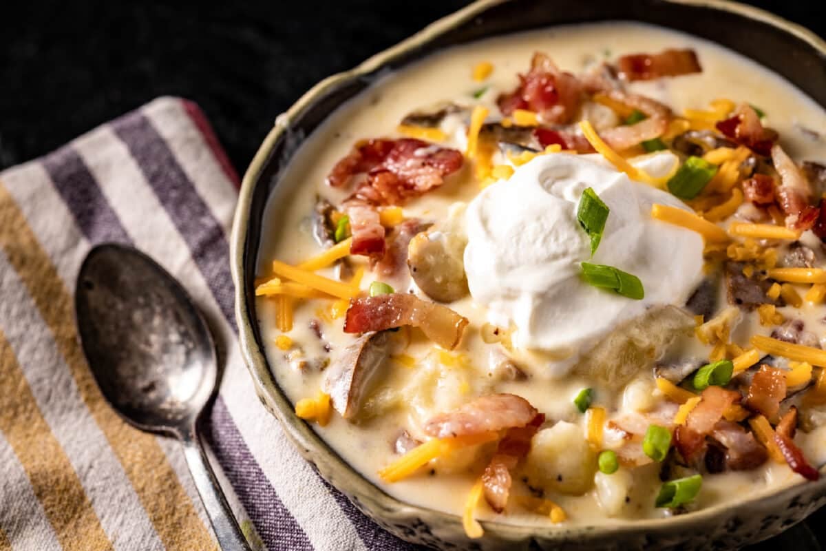 A bowl of baked potato soup topped with shredded cheese, bacon bits, and a dollop of sour cream next to a spoon on a cloth napkin.
