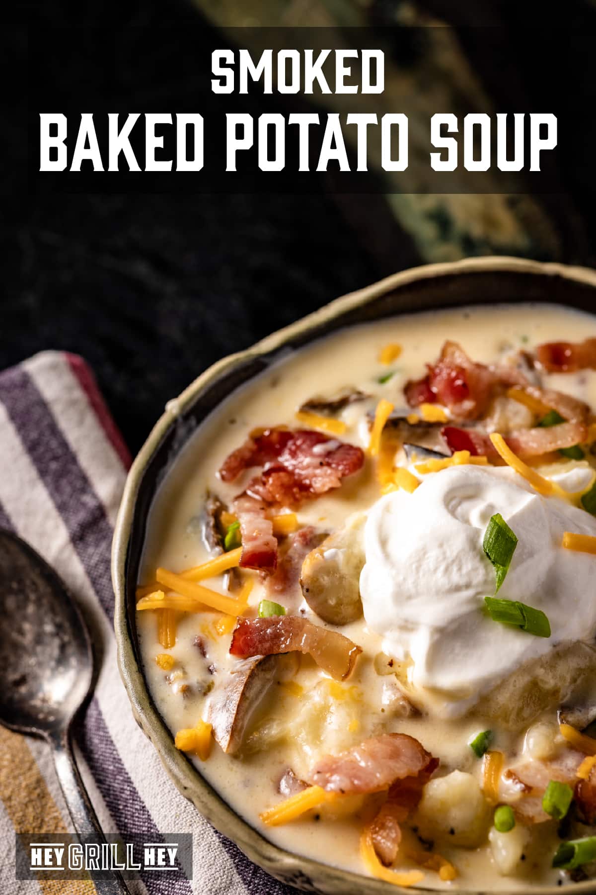 A closeup shot of a bowl of soup topped with shredded cheese, bacon bits, and a dollop of sour cream. The text overlay reads "Smoked Baked Potato Soup" at the top, and "Hey Grill Hey" at the bottom.
