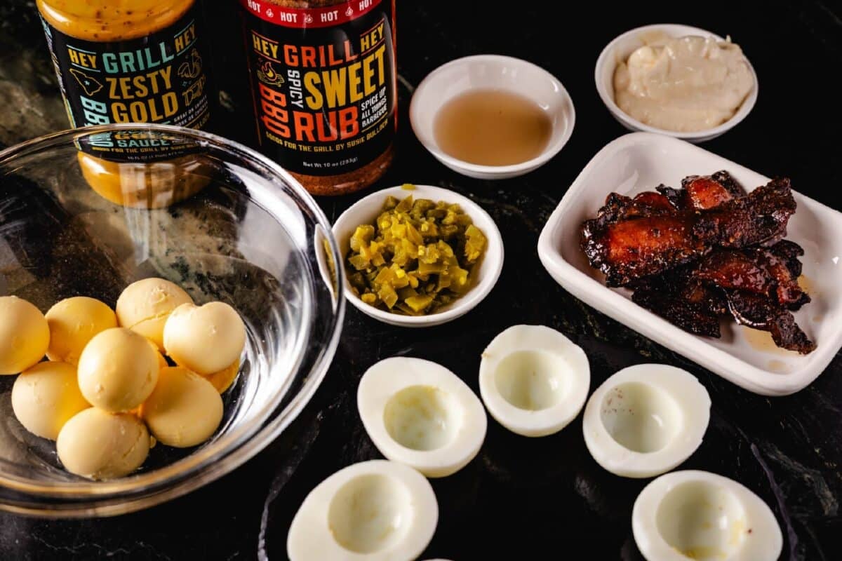 A clear bowl with cooked egg yolks, a black plate with cooked egg whites, a white plate of cooked bacon, white bowls with various ingredients, and bottles of Hey Grill Hey Zesty Gold BBQ Sauce and Hey Grill Hey Sweet Rub.