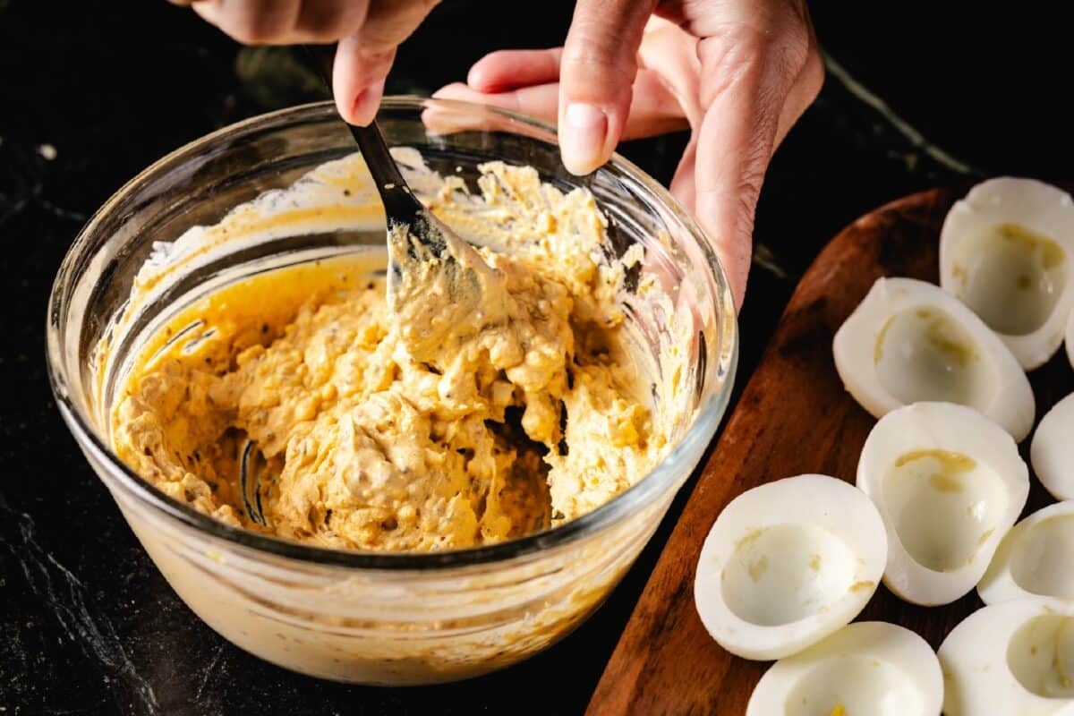 A chef's hand stirring an egg misture in a clear bowl, and a wooden tray of cooked egg whites.