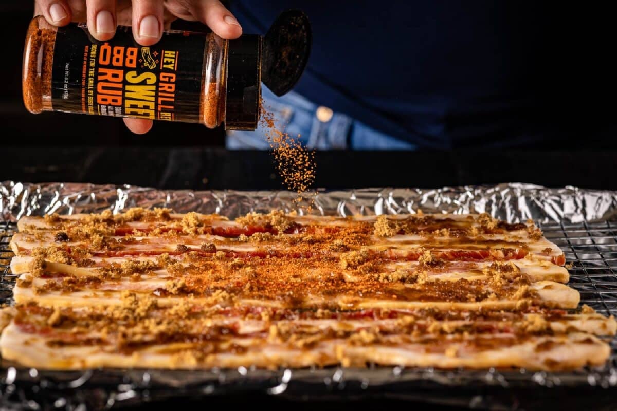 A wire rack topped with uncooked bacon above a foil-lined baking sheet, and a chef's hand sprinkling the bacon with Hey Grill Hey Sweet Rub.