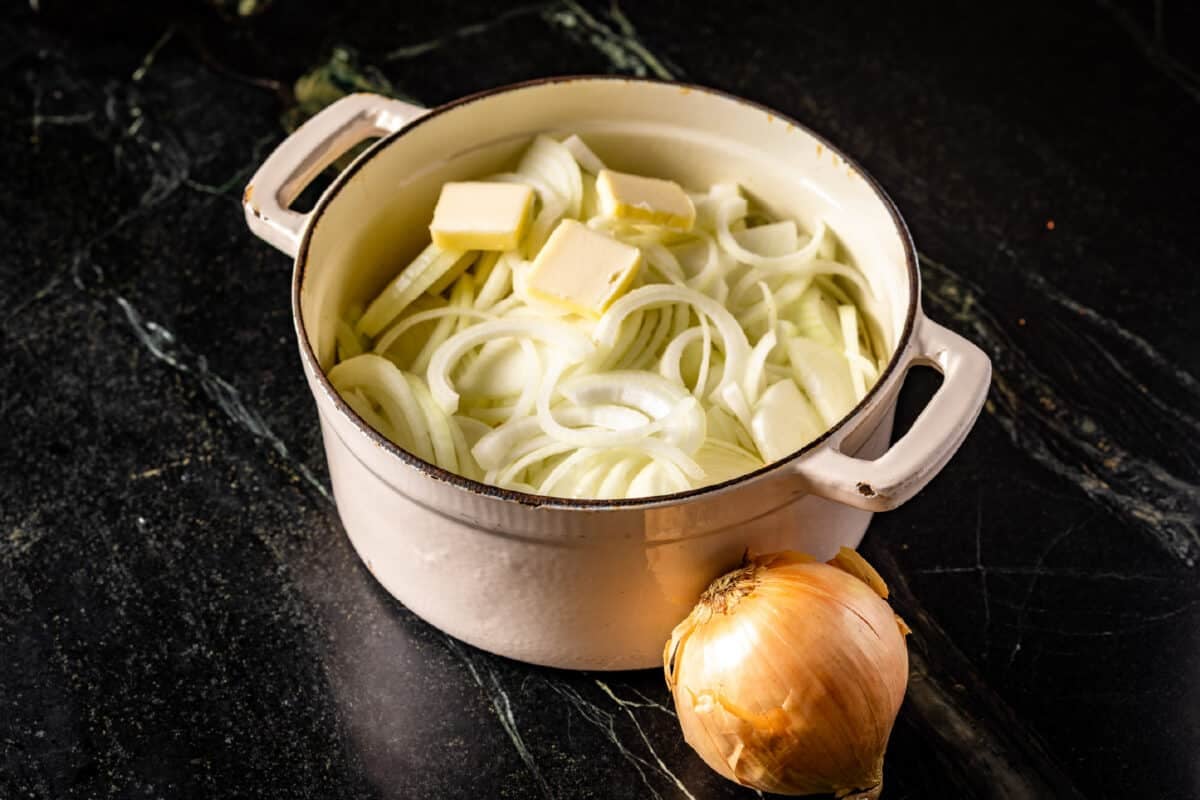 A white pot filled with sliced onions and pats of butter, and a whole onion, on a marble countertop.