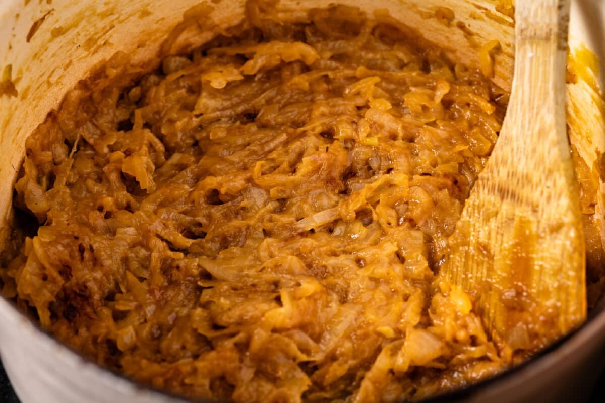 A wooden spoon stirring cooked onions and sauce in a white pot.