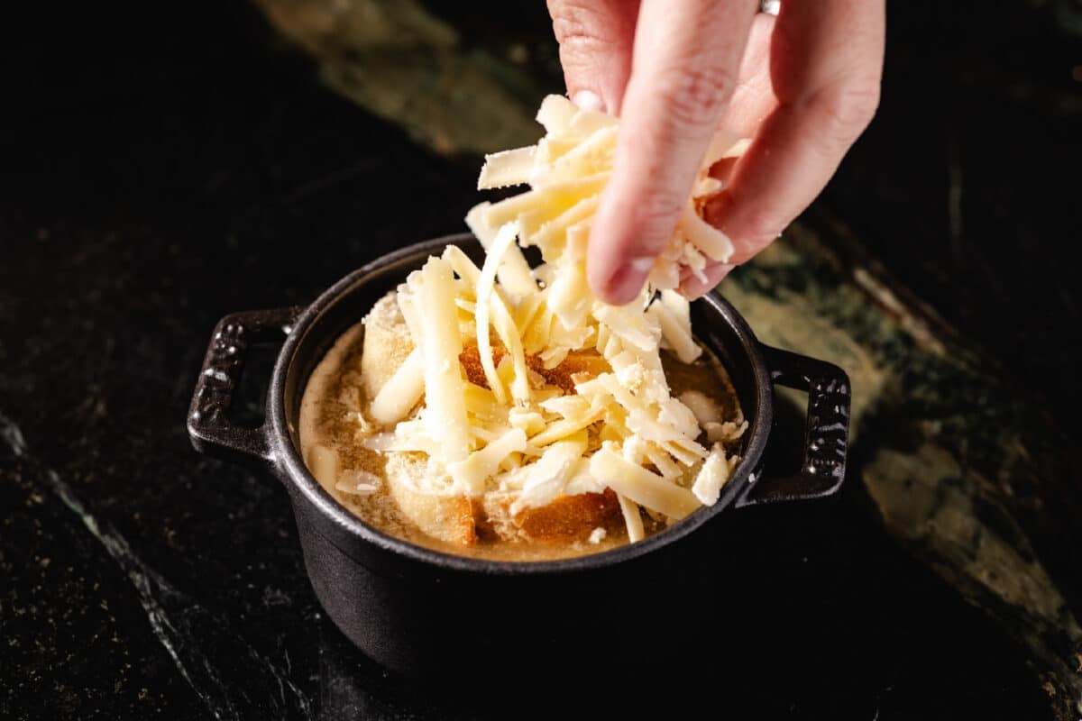 A chef's hands sprinkling shredded cheese on top of a soup in a cast iron pot.