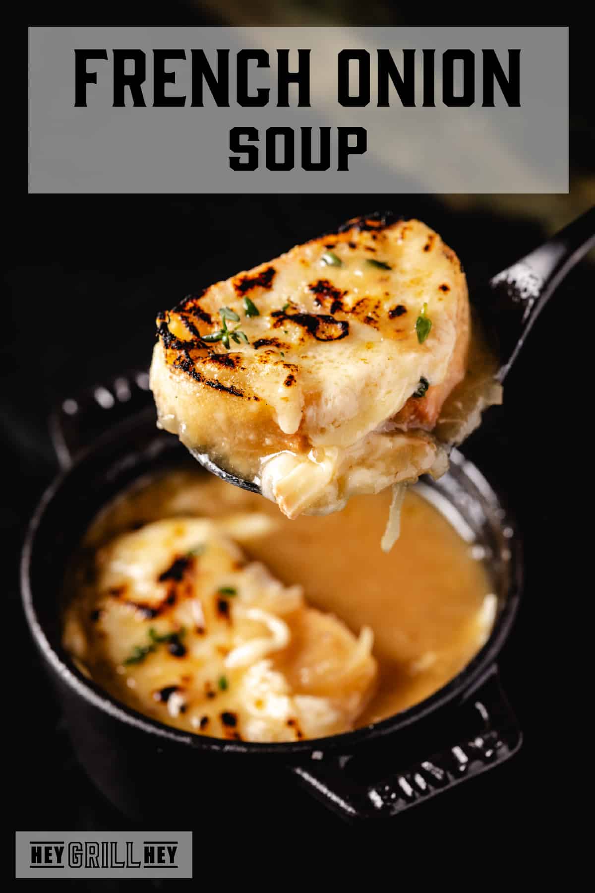 A spoon holding a baguette slice and crisp cheese above a cast iron bowl of soup. The text overlay reads "French Onion Soup" at the top, and "Hey Grill Hey" at the bottom.