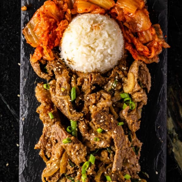 Beef bulgogi on a black, rectangular plate with a scoop of white rice and kimchi.