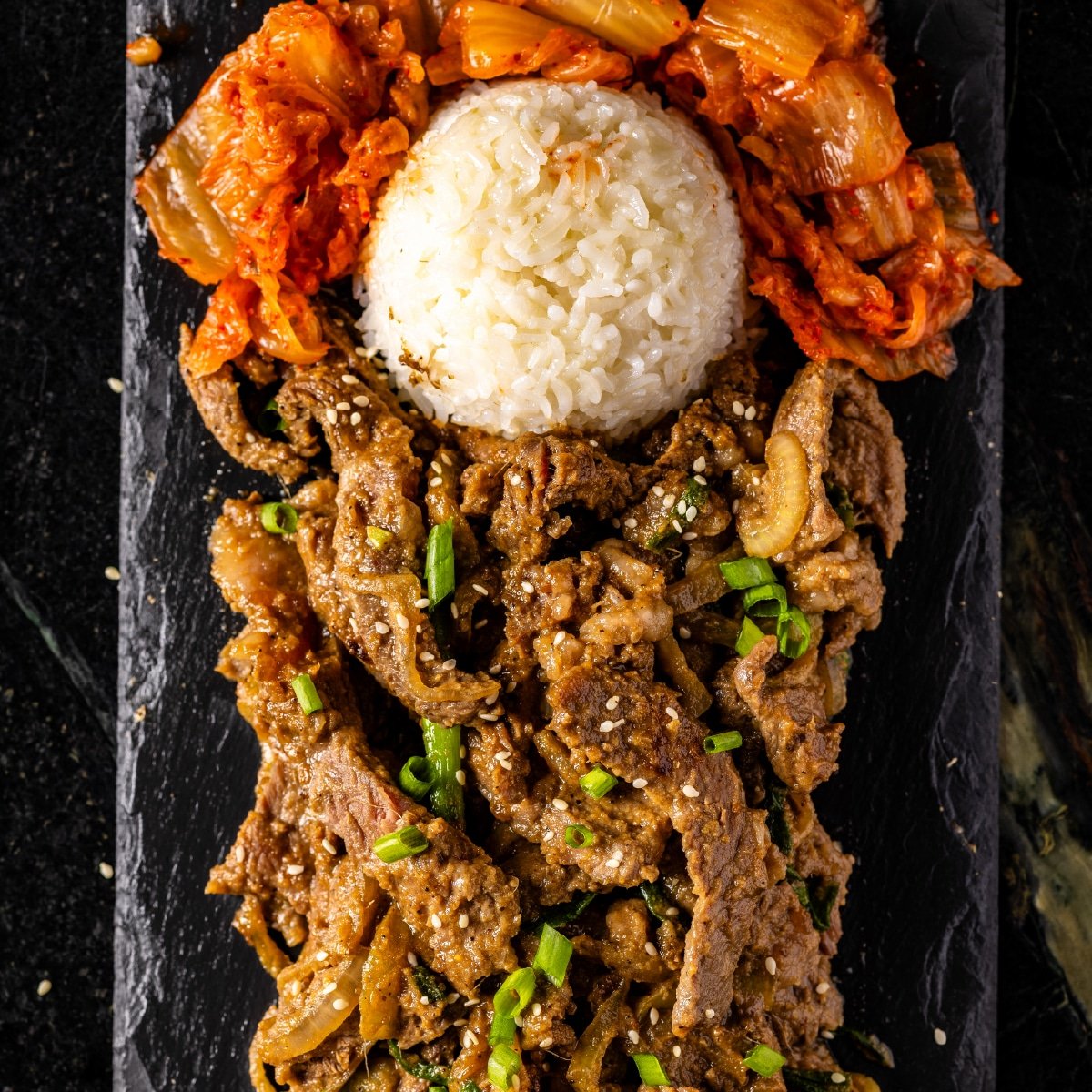 Beef bulgogi on a black, rectangular plate with a scoop of white rice and kimchi.