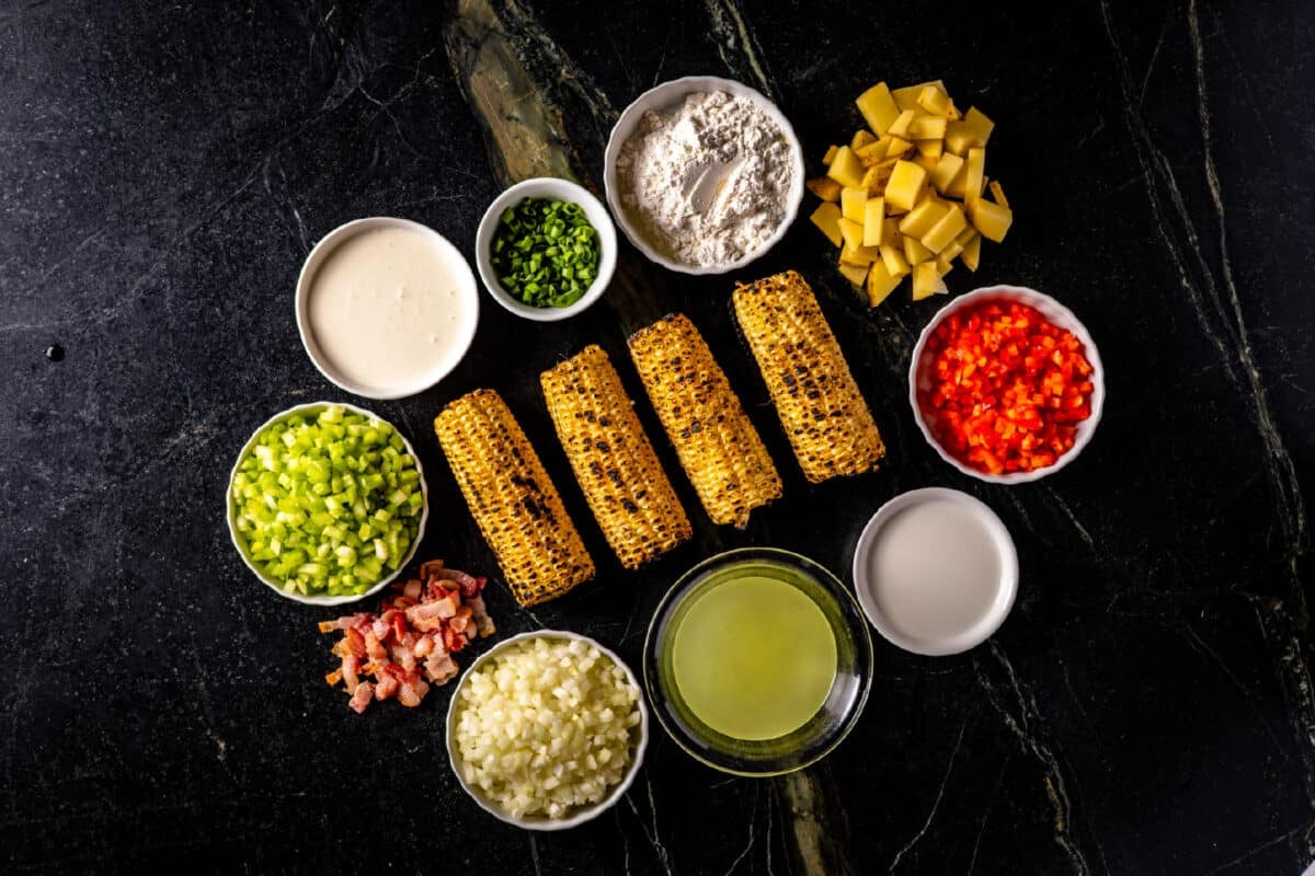 Four ears of grilled corn resting on a marble surface encircled by bowls of liquid ingredients and flour, and loose chopped ingredients.