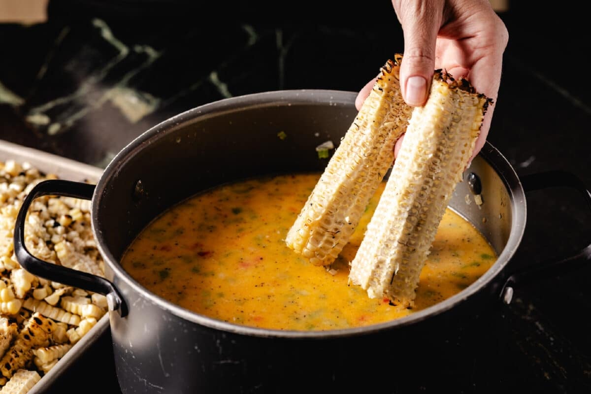 A chef's hand placing the cores of two cobs into a large pot of broth.