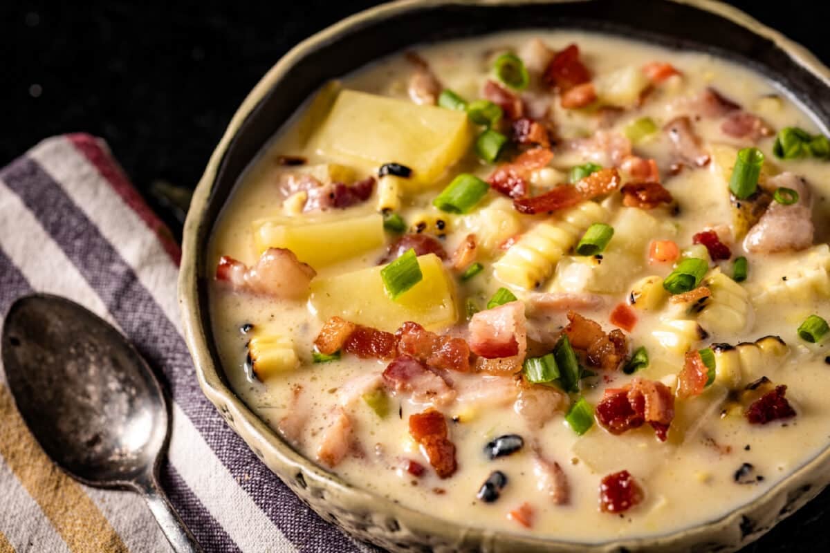 A bowl of corn, potato, and bacon soup, and a spoon resting on a striped, cloth napkin.