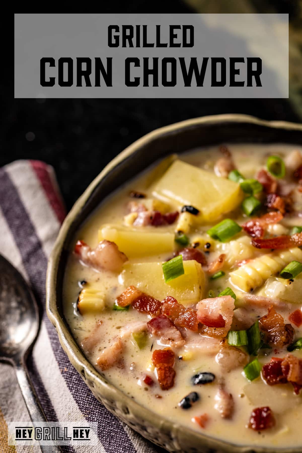 A bowl of corn, potato, and bacon soup, and a spoon resting on a striped, cloth napkin. The text overlay reads "Grilled Corn Chowder" at the top, and "Hey Grill Hey" at the bottom.