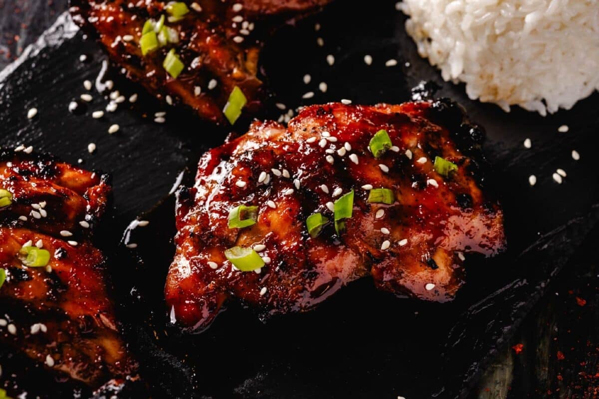Smoked chicken thighs topped with sliced green onions and sesame seeds, and a scoop of rice on a black platter.