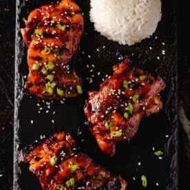 Smoked chicken thighs topped with sliced green onions and sesame seeds, and a scoop of rice on a black platter.