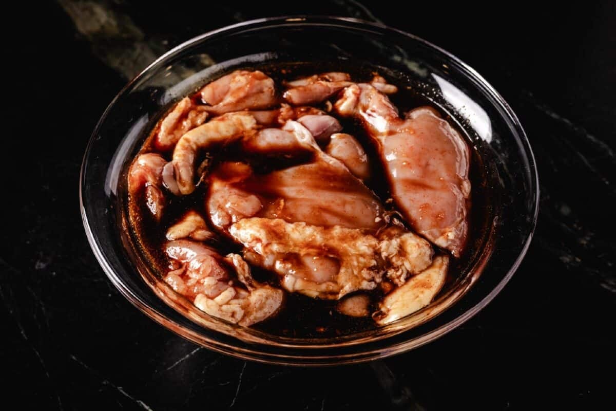 A clear bowl of poultry and marinade sitting on a marble surface.