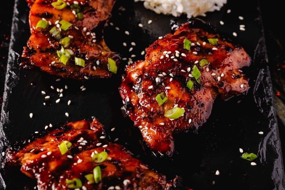 Smoked chicken thighs topped with sliced green onions and sesame seeds, and a scoop of rice on a black platter.