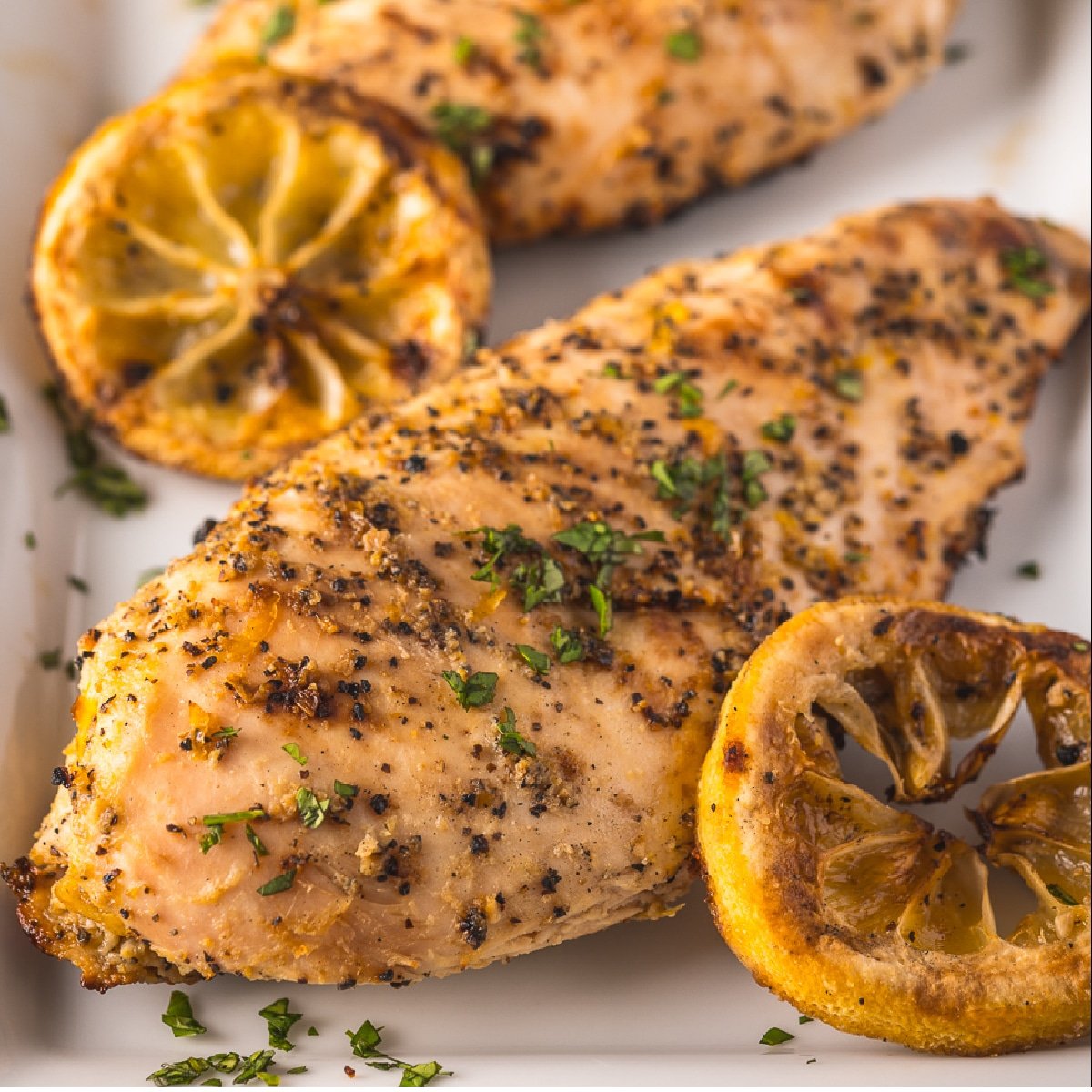 Two grilled lemon pepper chicken breasts on a platter next to grilled lemon slices.