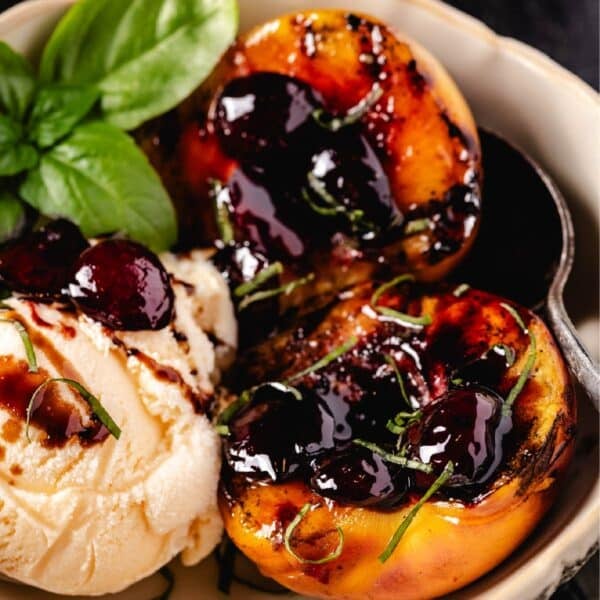 A bowl with peach halves and a scoop of vanilla ice cream topped with a cherry glaze, and a mint garnish.