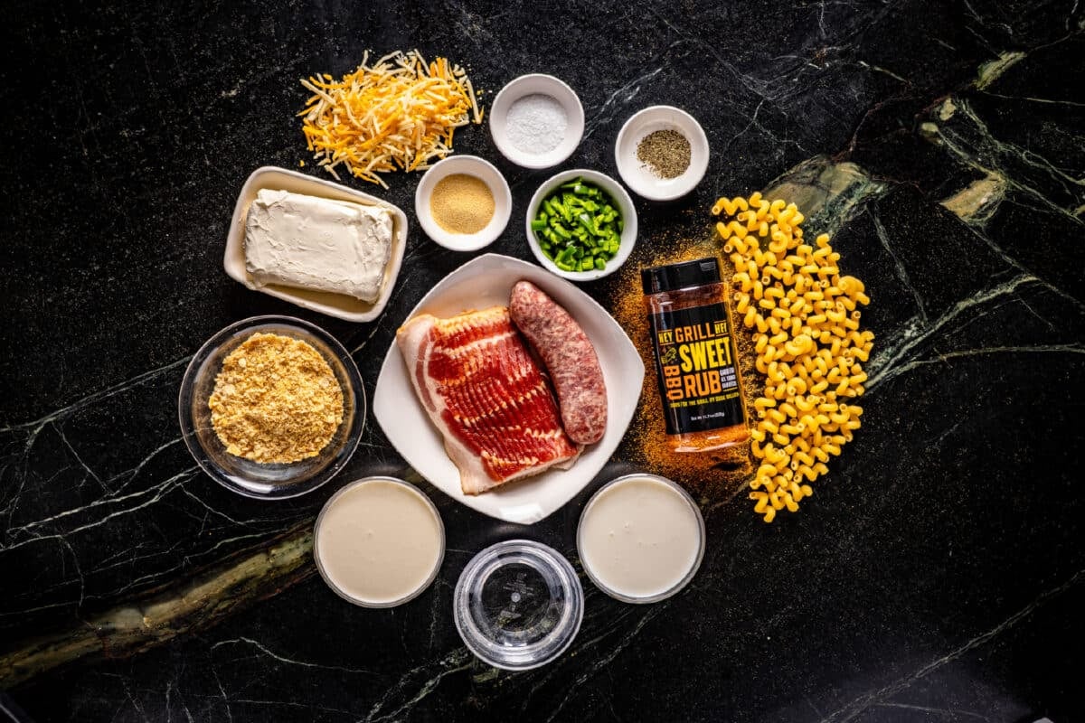 An overhead shot of a white plate with raw bacon and sausage, a bottle of Hey Grill Hey Sweet Rub, loose corn kernels and shredded cheese, and several bowls of raw ingredients on a marble surface.