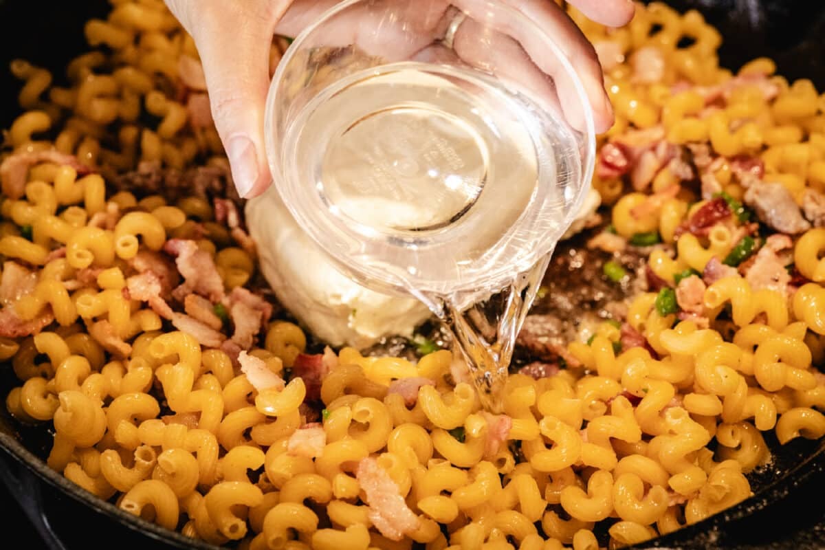 A chef's hands pouring a clear liquid from a clear bowl into a skillet filled with pasta noodles.