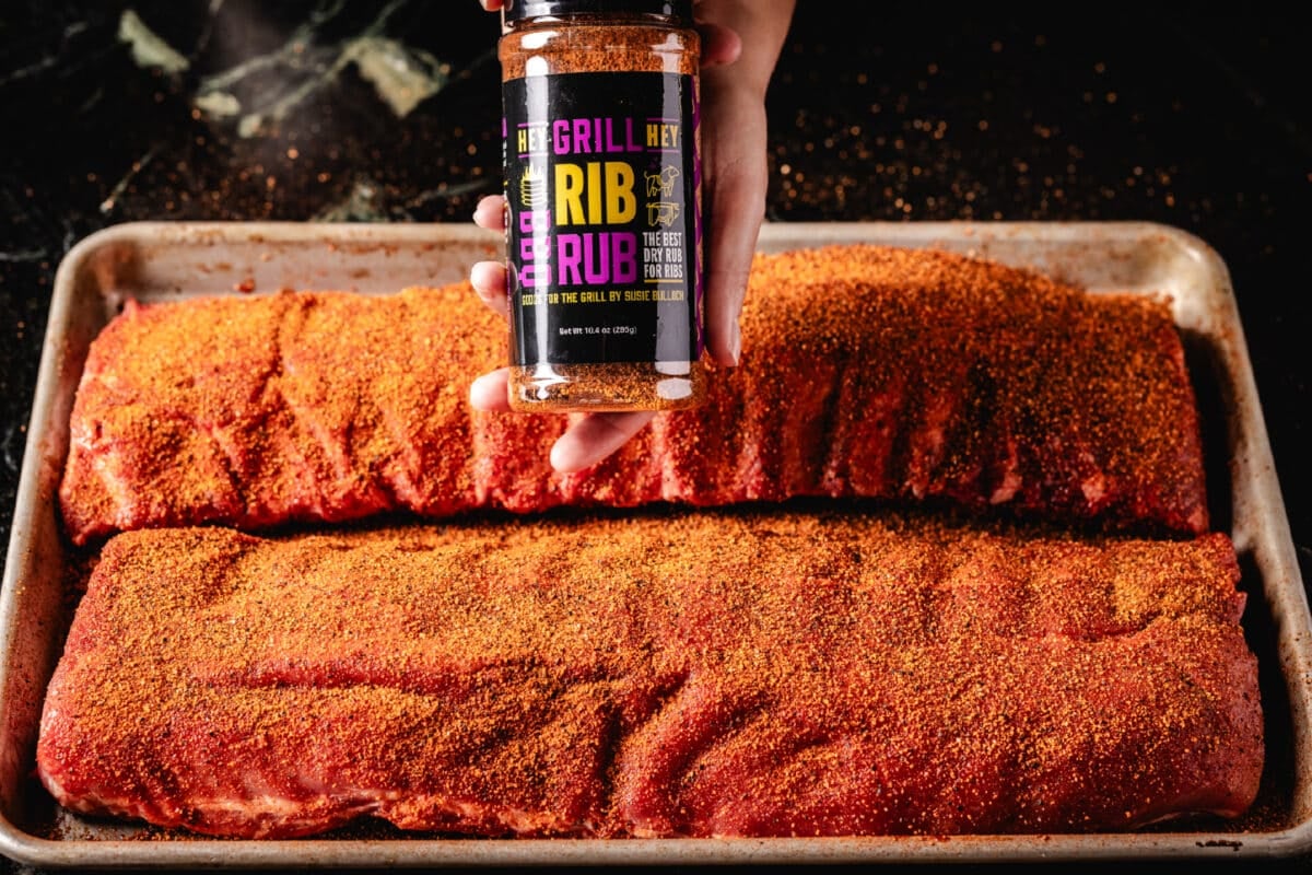 A chef's hand holding a bottle of Hey Grill Hey Rib Rub above two seasoned racks of ribs resting on a baking sheet.