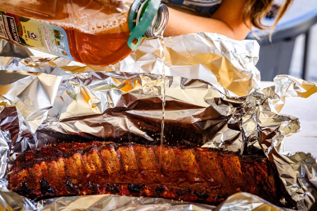 A jug of apple juice being poured into a foil packet containing a seasoned rack of ribs.